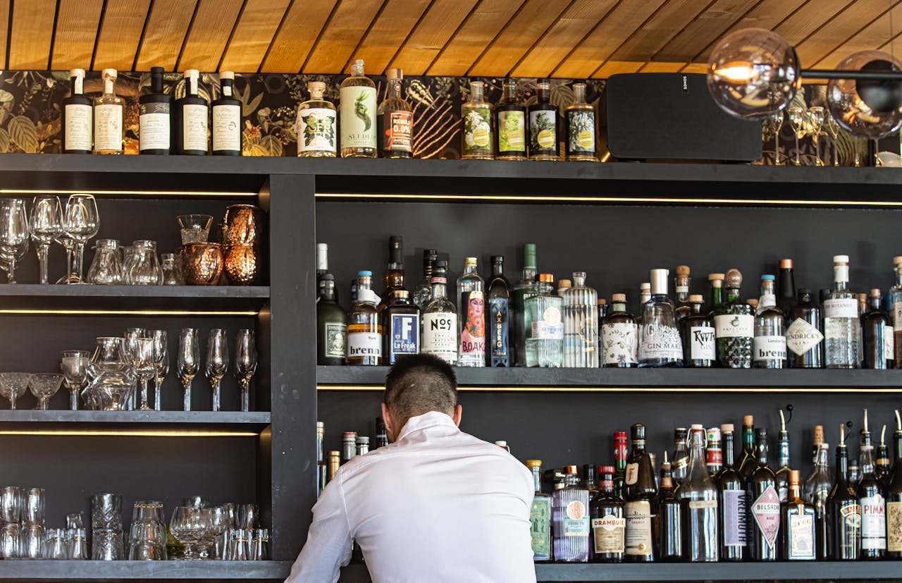 Bartender in white shirt arranging various liquor bottles on shelves in a stylish bar setting.